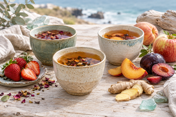 Three ceramic cups of tea surrounded by strawberries, peach, plums, apples, and sliced ginger on a wooden table overlooking the ocean.