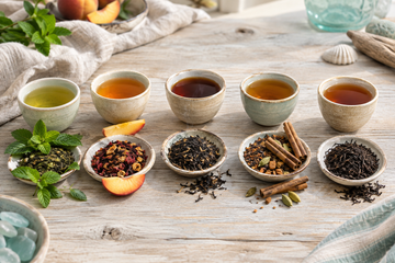 Five ceramic tasting cups beside dishes of loose tea, fresh mint, peach slices, and cinnamon sticks on a wooden table.