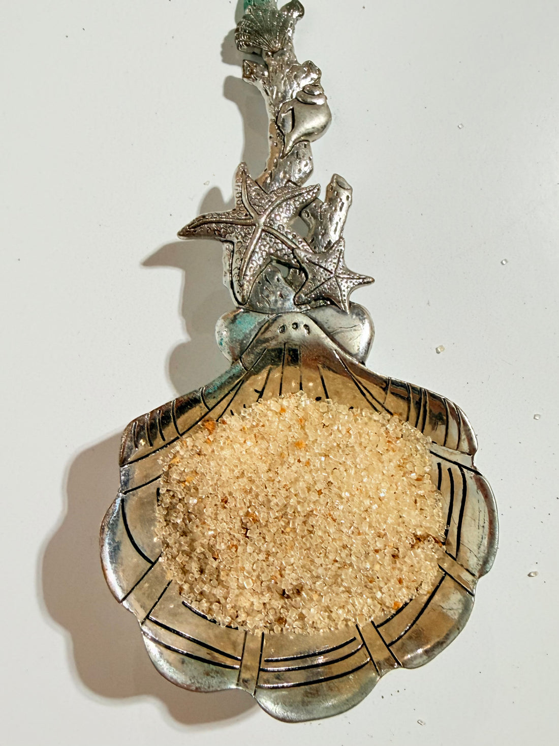 A silver-toned decorative spoon or scoop shaped like a scalloped shell, filled with coarse sand, shown against a plain off-white background.