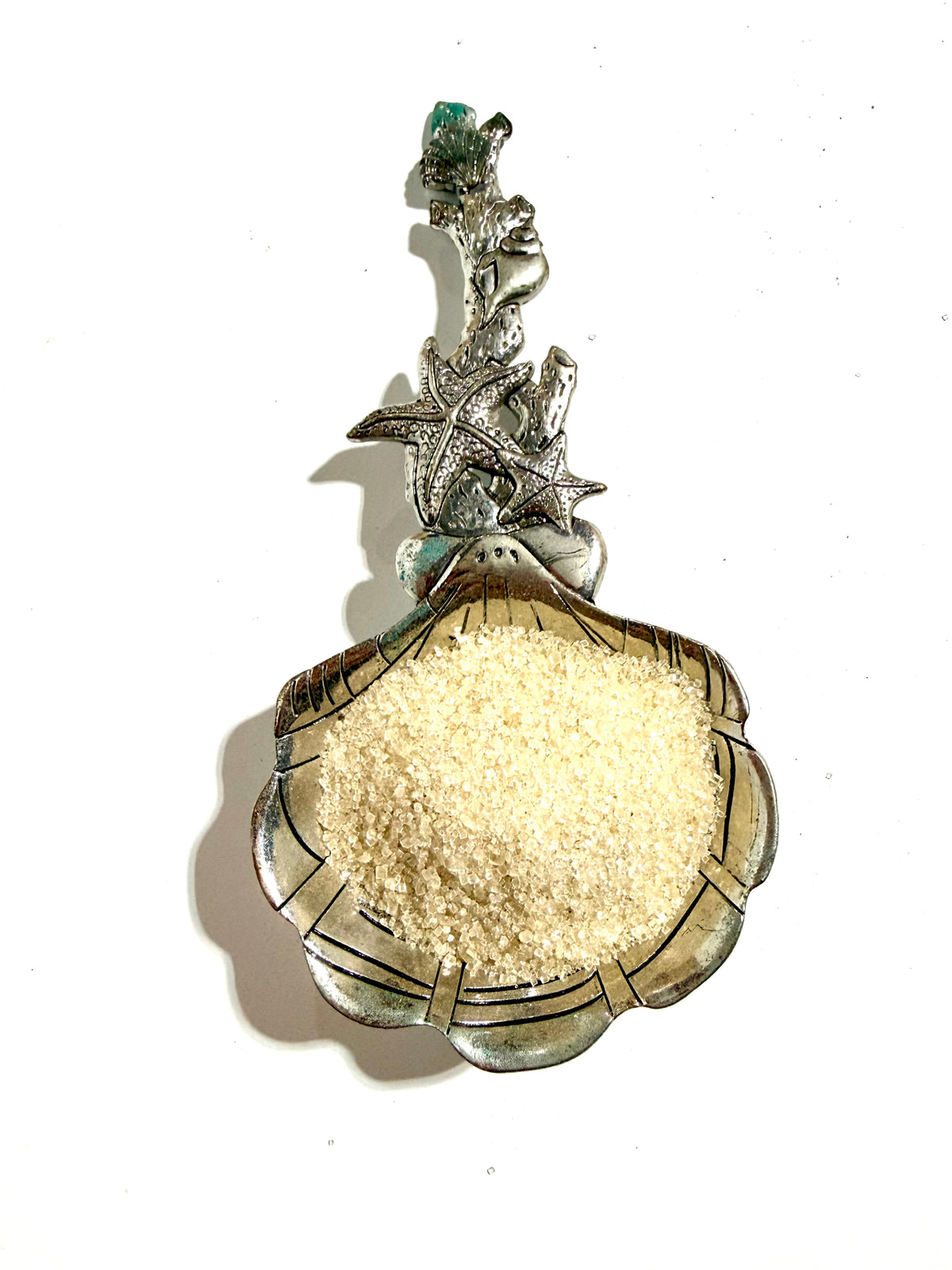 Lime Basil infused sugar in a silver-toned shell-shaped spoon with decorative sea-life details on white