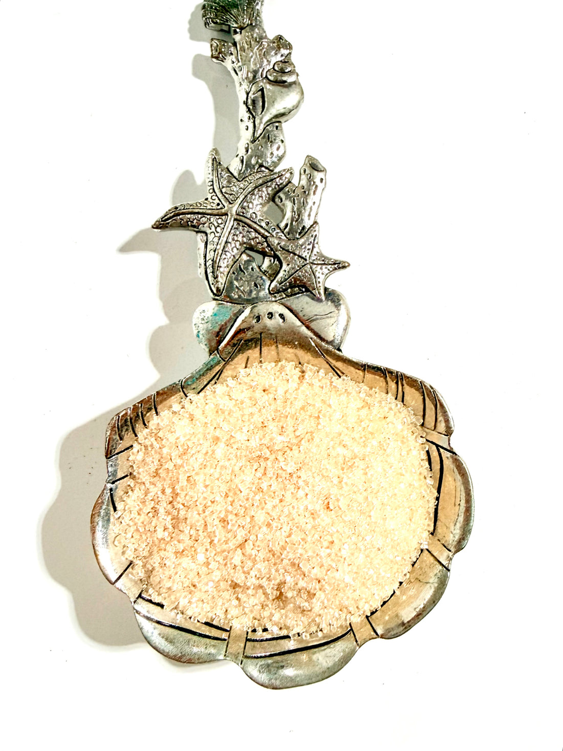 A silver-toned seashell-shaped bowl filled with coarse sea salt, featuring a starfish and coral design on the handle, set against a plain white background.