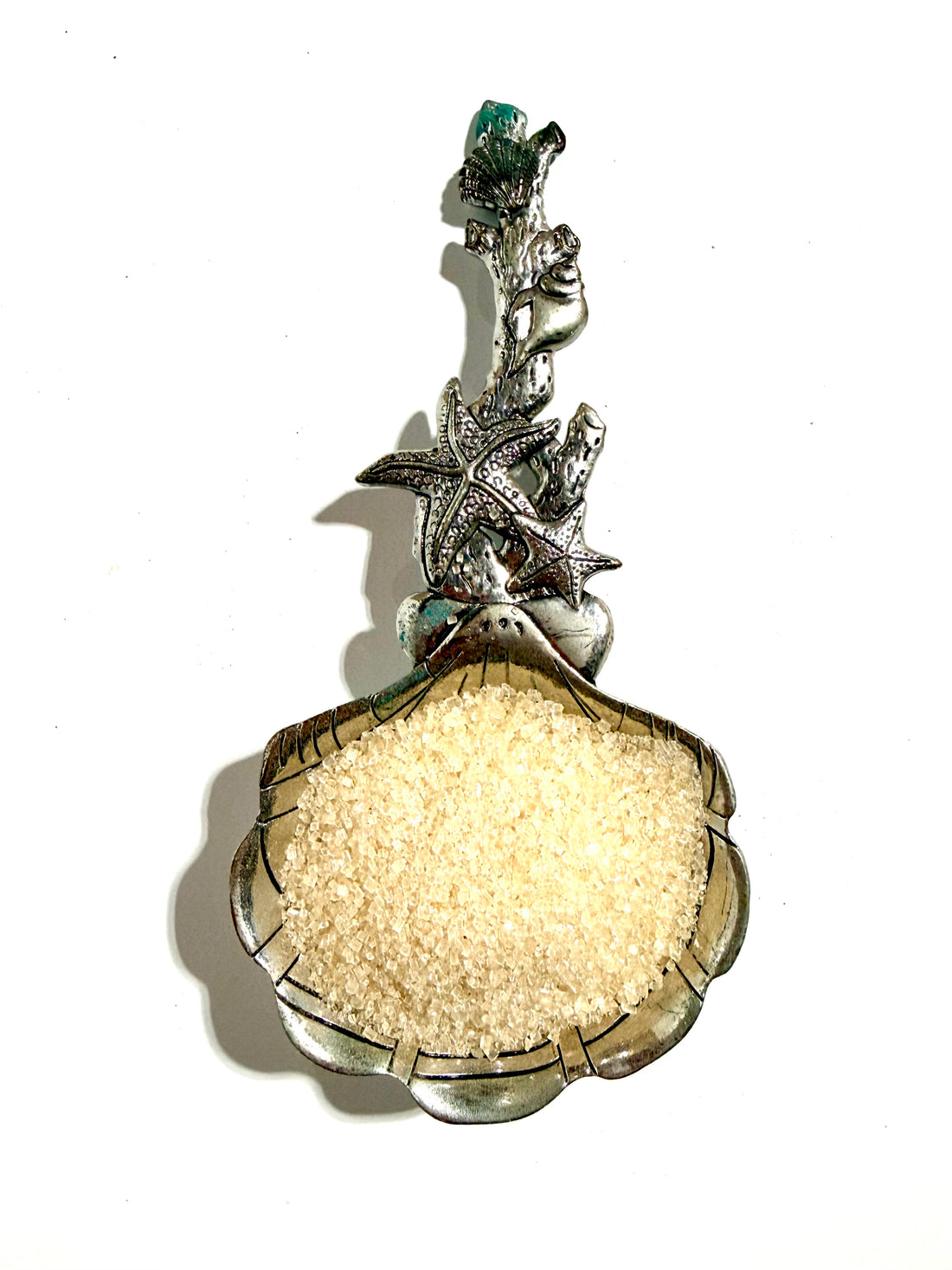 A silver-toned decorative spoon or scoop shaped like a clam shell, filled with coarse white sugar, photographed against a plain white background.