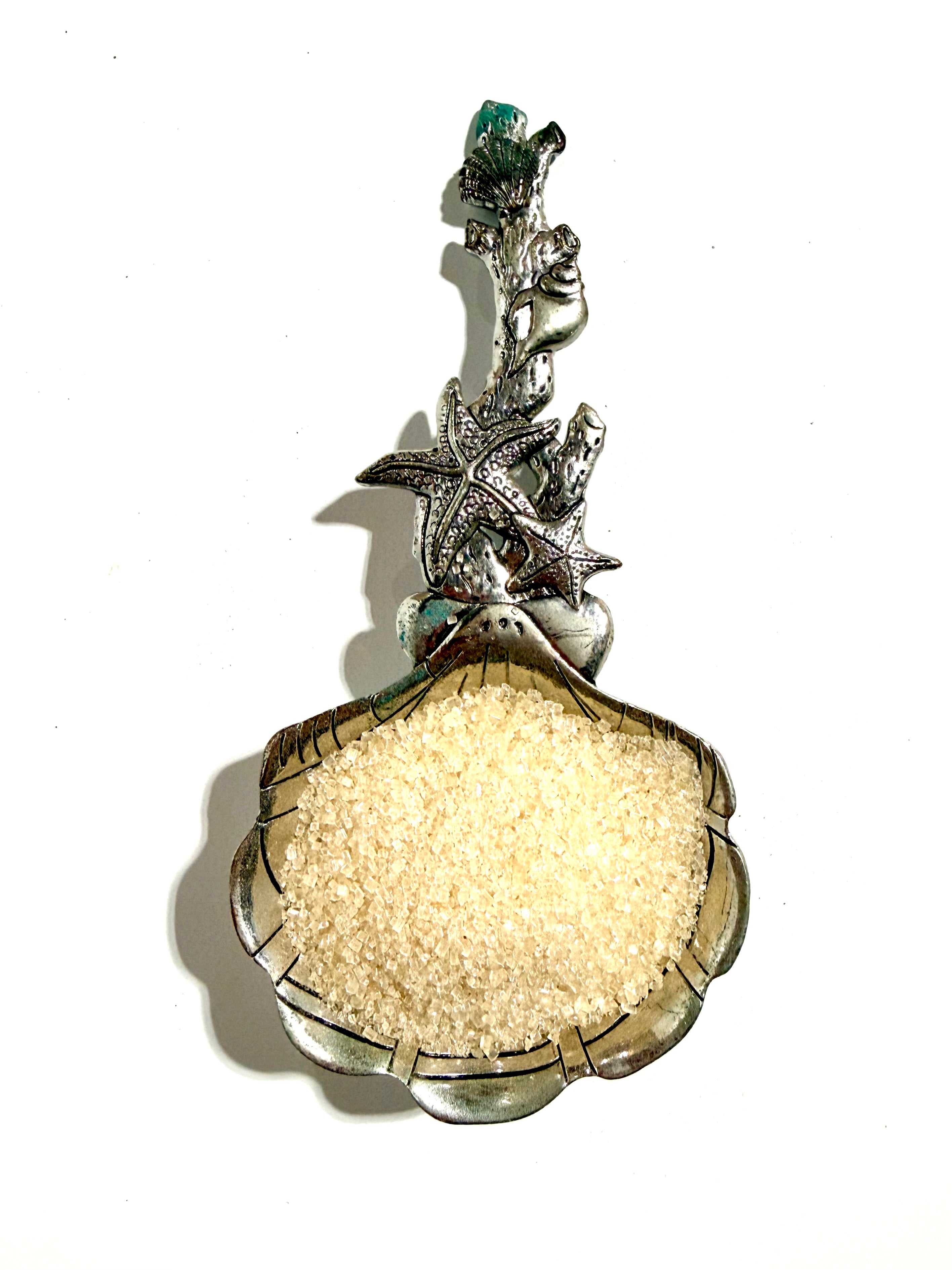A silver-toned decorative spoon or scoop shaped like a clam shell, filled with coarse white sugar, photographed against a plain white background.