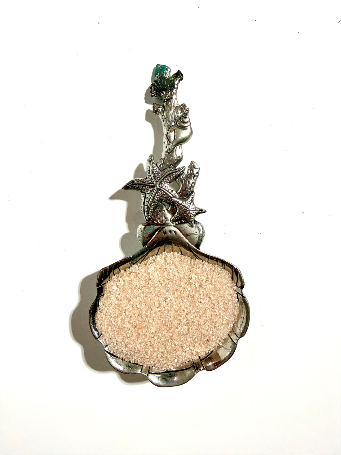 A silver-toned seashell-shaped jewelry box filled with light beige sand, featuring starfish and coral details on the clasp, set against a plain white background.