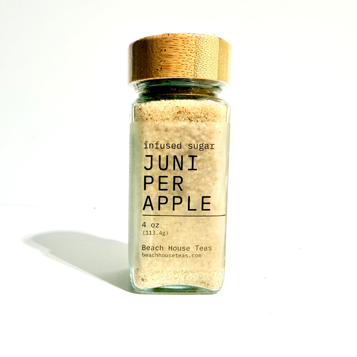 A clear glass bottle of Beach House Teas 'Juni Per Apple' infused sugar with a light wood cap, set against a plain white background with a soft shadow.