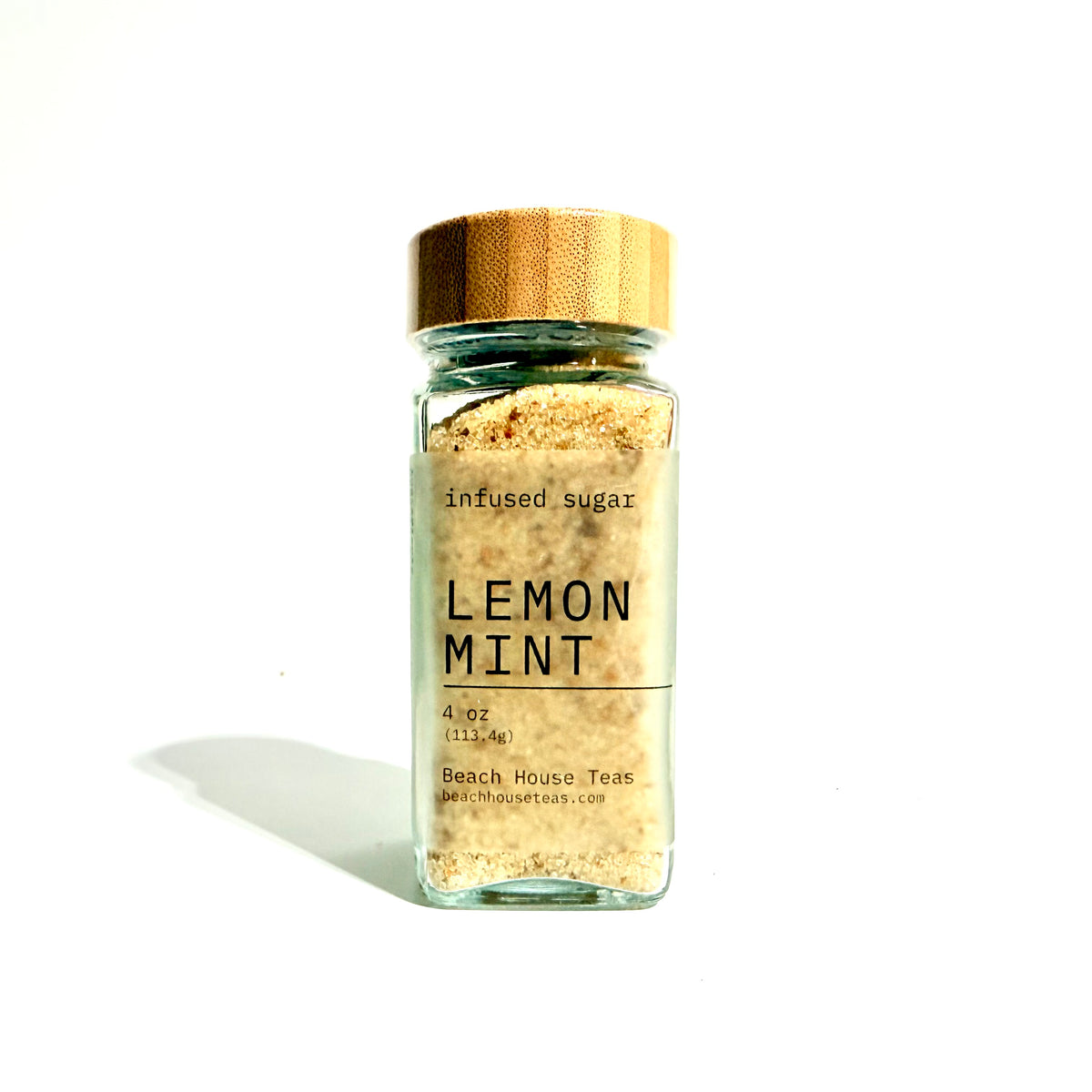 A small clear glass jar filled with light tan infused sugar, topped with a metallic gold cap, and labeled 'LEMON MINT' on a white background.
