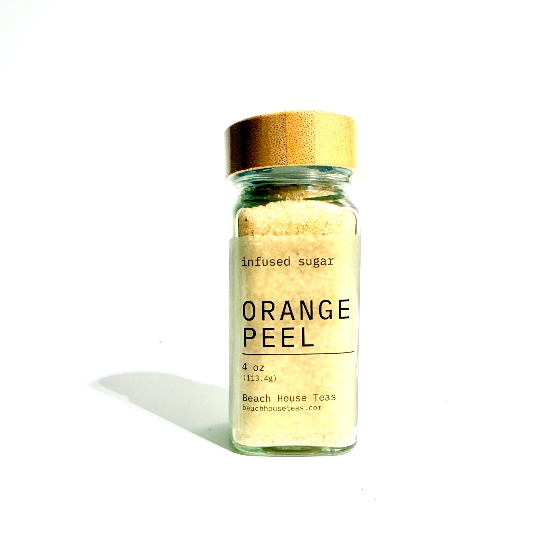 A small glass jar of Beach House Teas Orange Peel infused sugar, featuring a gold metallic cap and a cream-colored label with black text, set against a plain white background with a soft shadow.