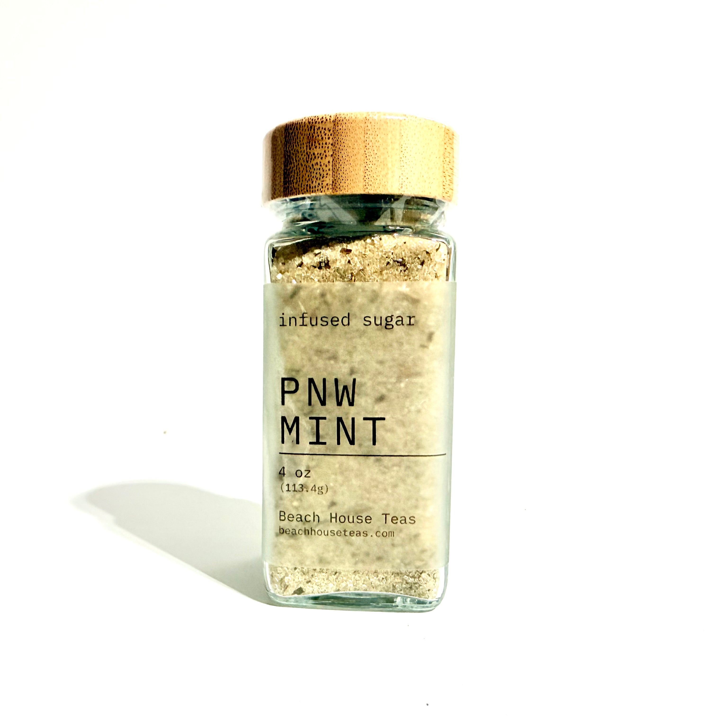 A clear glass jar of PNW Mint infused sugar with a metallic gold lid and a white label, set against a plain white background with a soft shadow.