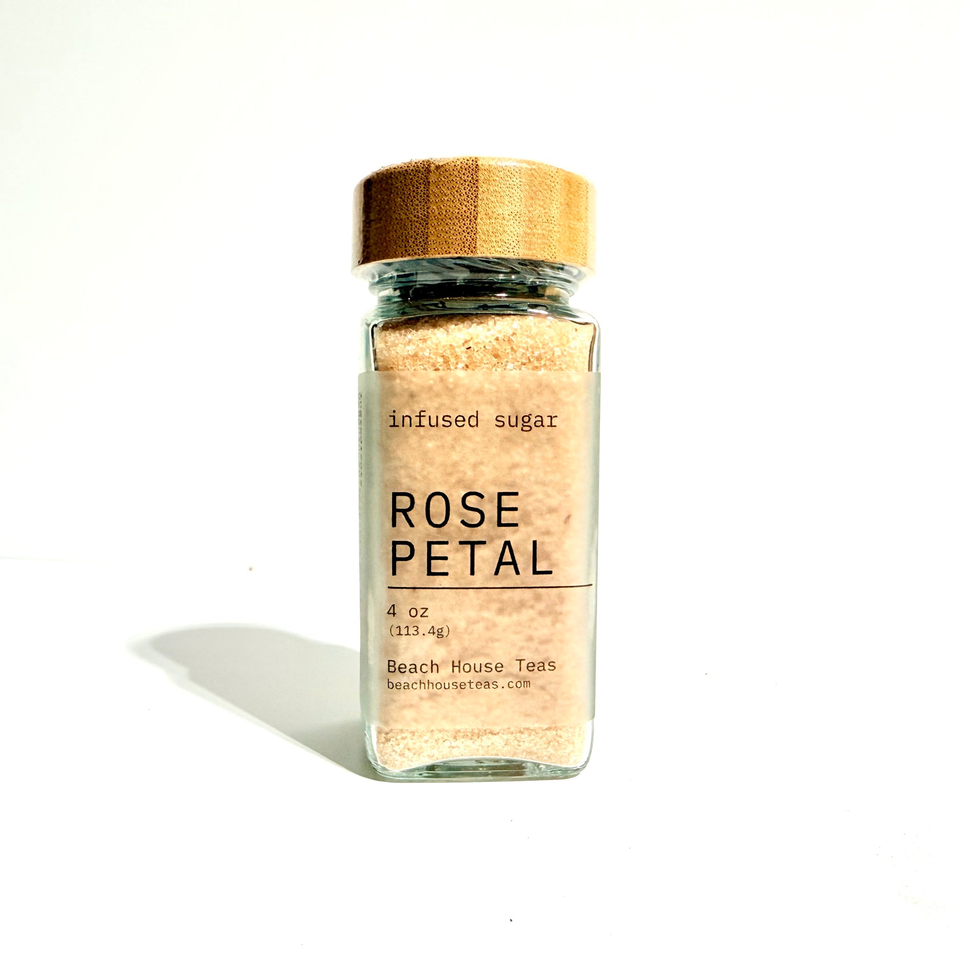 A clear glass jar filled with pale pink-beige infused sugar, topped with a natural wood lid, and labeled 'ROSE PETAL' on a white background.