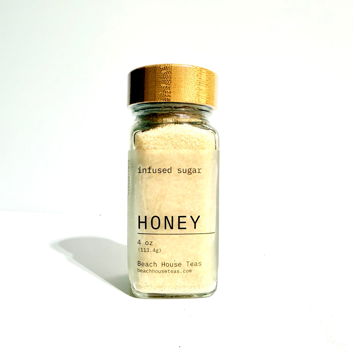 A small clear glass jar filled with pale honey-colored sugar, topped with a metallic gold lid, and labeled 'HONEY' with 'Beach House Teas' visible on the front, photographed on a white background with a soft shadow.