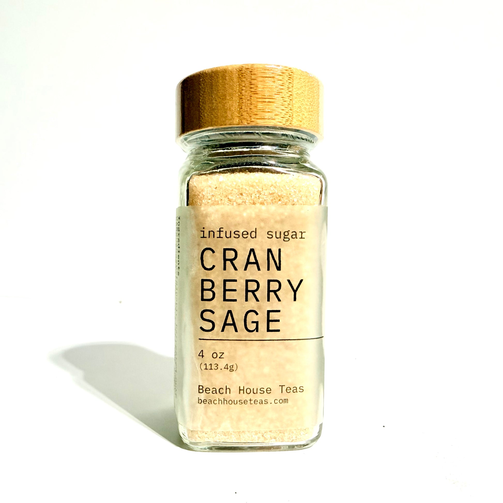 A small clear glass jar filled with light tan infused sugar, topped with a natural wood lid and a cream label reading 'infused sugar CRAN BERRY SAGE' and 'Beach House Teas'.