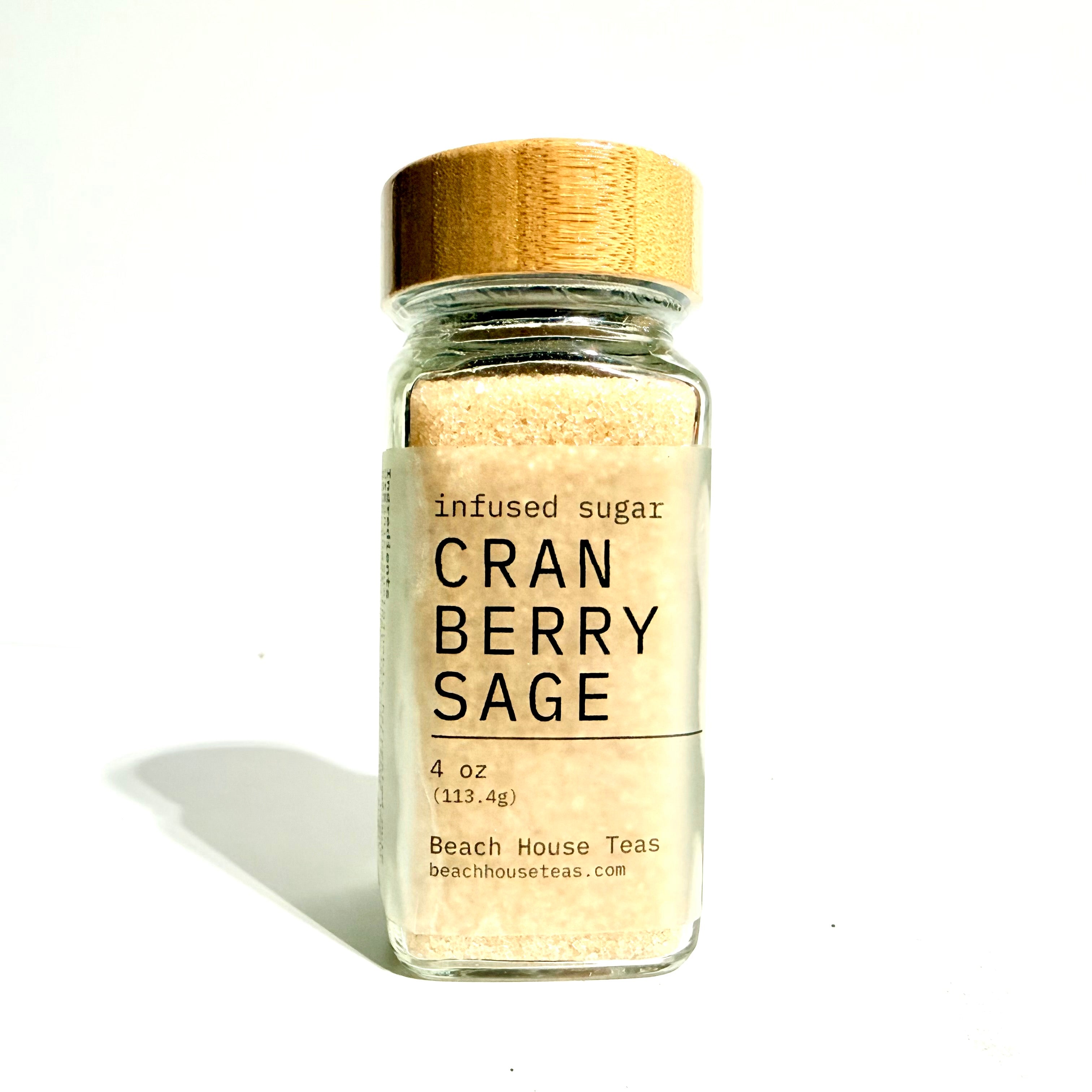 A small clear glass jar filled with light tan infused sugar, topped with a natural wood lid and a cream label reading 'infused sugar CRAN BERRY SAGE' and 'Beach House Teas'.