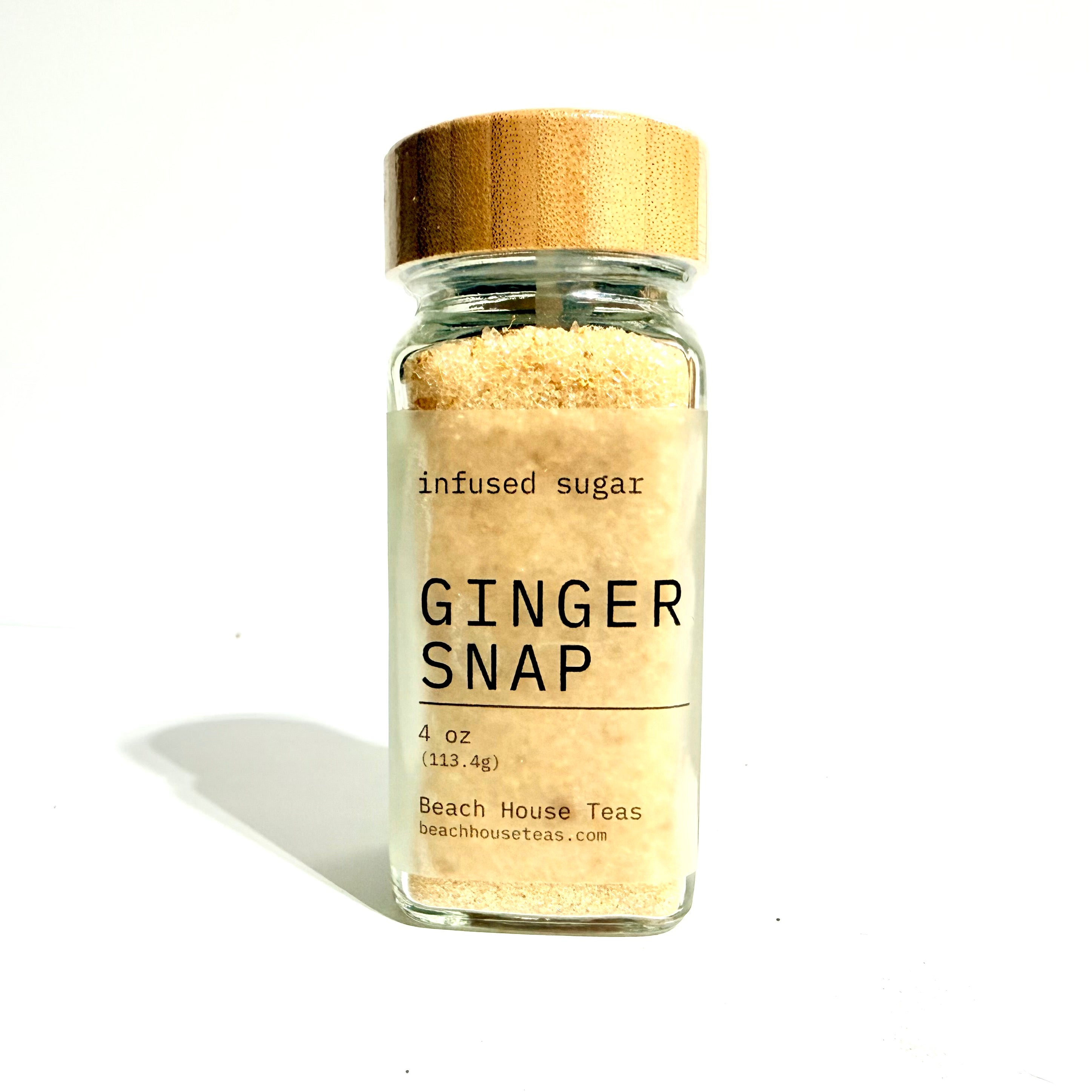 A clear glass jar filled with light tan infused sugar, topped with a bamboo-style gold lid, labeled 'GINGER SNAP' by Beach House Teas, on a white background with a soft shadow.