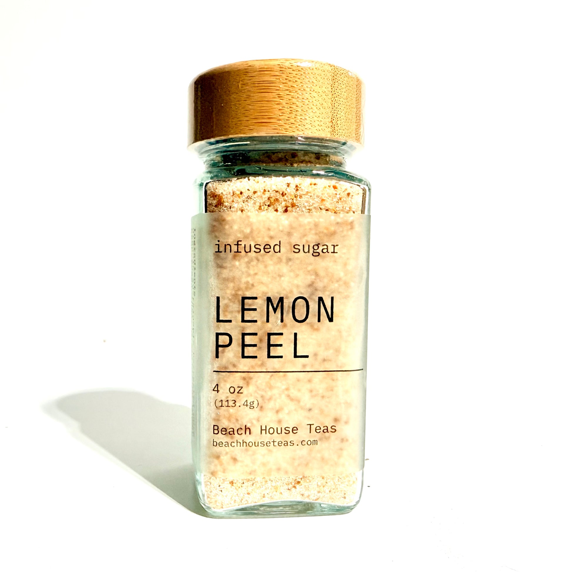 A clear glass jar filled with beige lemon peel infused sugar, featuring a light wood-grain lid and a white label with black text that reads 'LEMON PEEL' and 'Beach House Teas'.