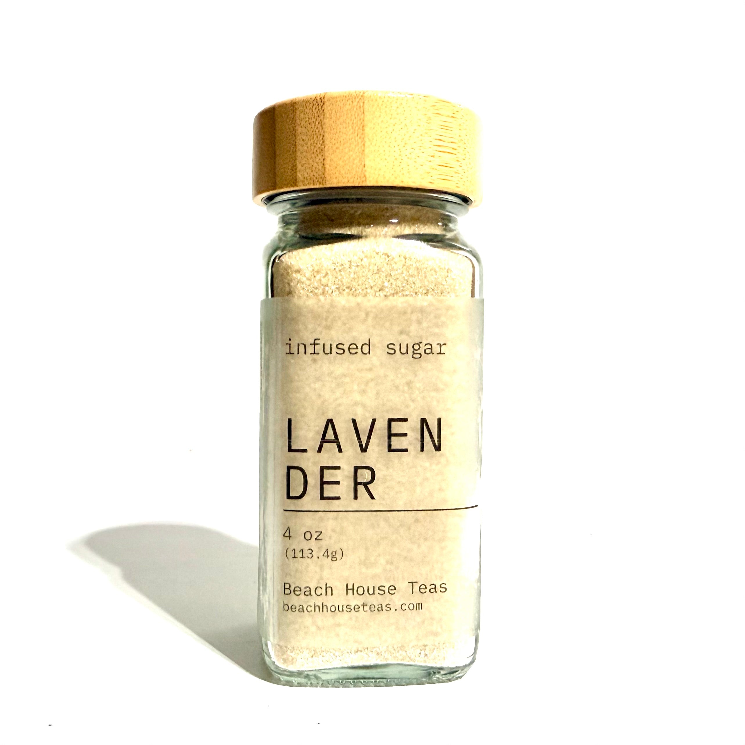 A small clear glass jar filled with pale beige infused sugar, topped with a light wood lid, and labeled 'LAVENDER' with 'infused sugar' above it. The jar is photographed on a white background with a soft shadow to the left.