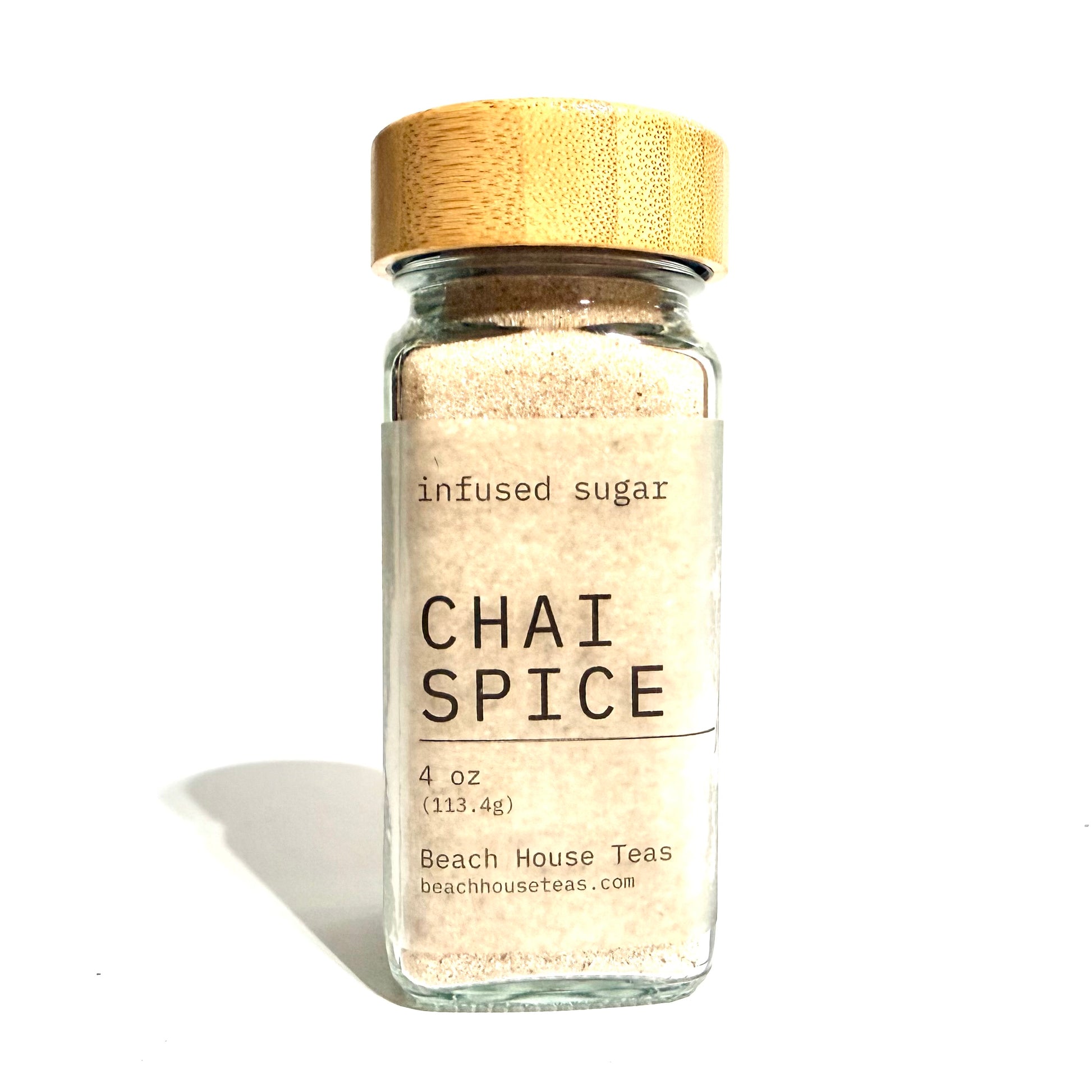 A clear glass jar filled with light beige infused sugar, featuring a natural bamboo lid and a white label that reads 'infused sugar CHAI SPICE 4 oz Beach House Teas'.