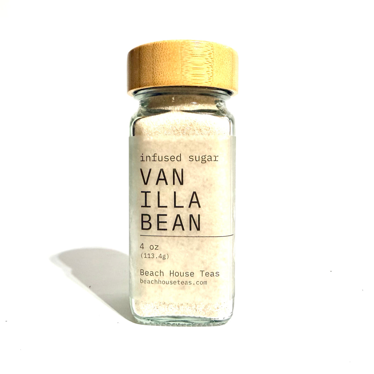 A small clear glass jar filled with a light beige powder, topped with a natural wood cap, and labeled 'infused sugar VAN ILLA BEAN' with 'Beach House Teas' at the bottom, photographed on a white background with a soft shadow.