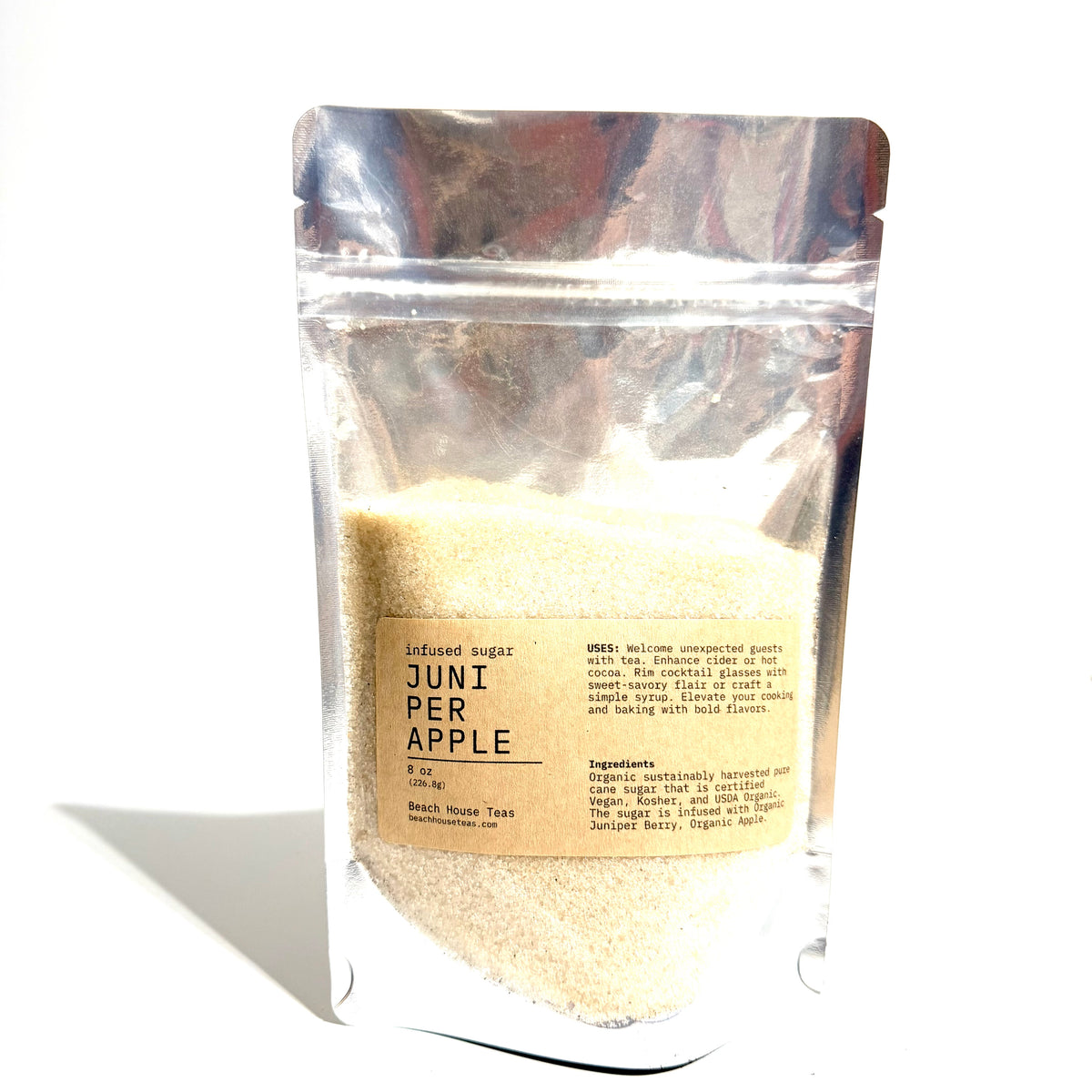 A stand-up resealable pouch of infused sugar labeled 'JUNI PER APPLE' by Beach Breeze Tea, filled with pale beige sugar crystals, photographed on a white background with a cast shadow.