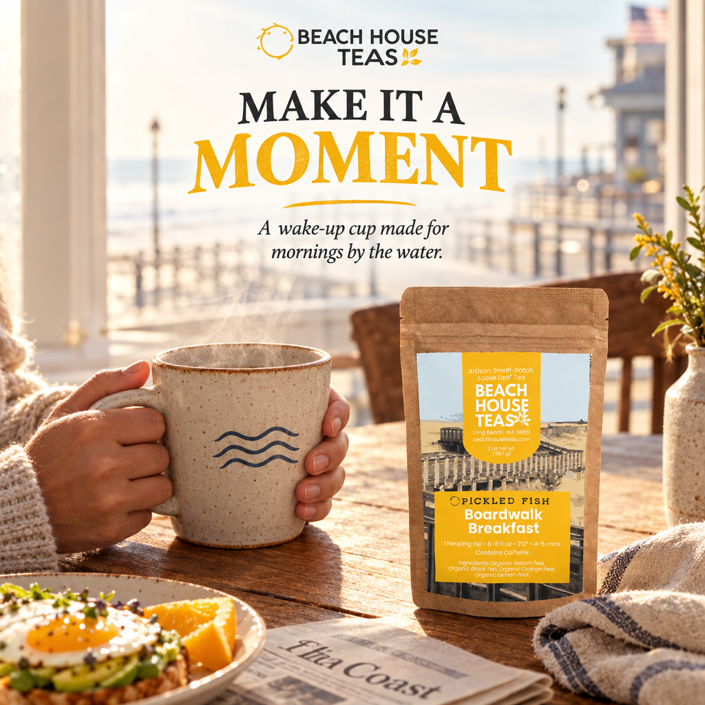 Hands holding a ceramic tea mug beside a Boardwalk Breakfast pouch, breakfast and newspaper by a coastal window.